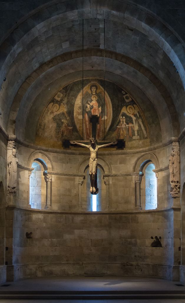 A rounded stone recess with three narrow arched windows and a painted half-dome is framed by a barrel-vaulted bay, with a large crucifix hanging in front.