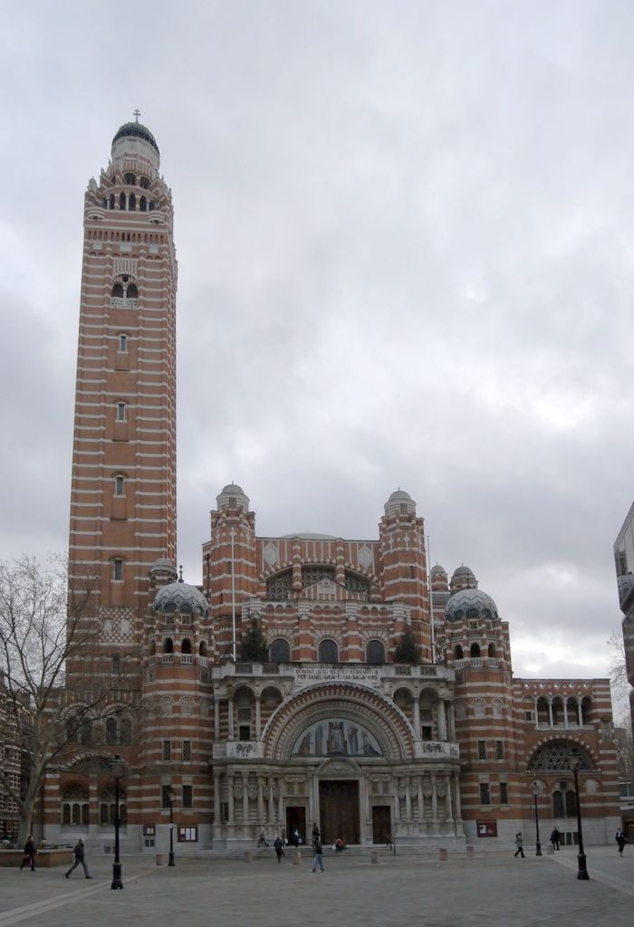 A tall striped brick-and-stone tower with a domed crown and cross stands beside the cathedral’s broad arched entrance and forecourt.