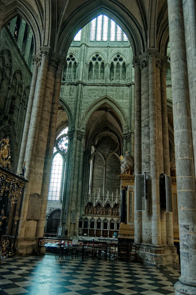A tiled passage runs between tall clustered piers and a screen into a vaulted transept with windows, chairs and barriers.