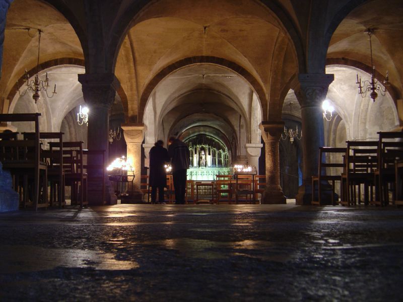 Low stone vaults and rounded arches recede between thick columns toward an illuminated altar area, with chairs and two visitors in the centre.