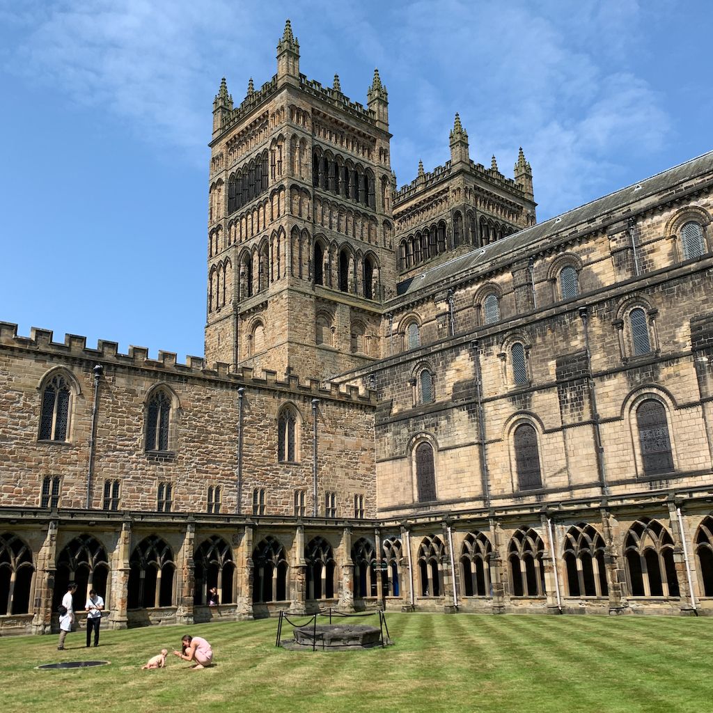 A grass court is bordered by low traceried cloister walks and taller cathedral walls, with towers above and visitors below.