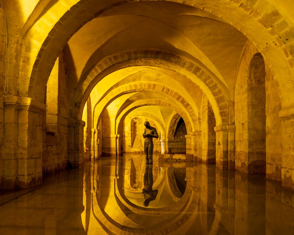 Water covers the crypt floor and reflects round arches, vaults and a standing modern sculpture under yellow lighting.
