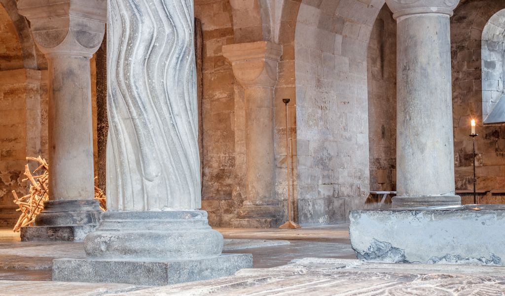 Plain cylindrical and undulating carved stone shafts rise from different bases beside paving, a small window and candles.