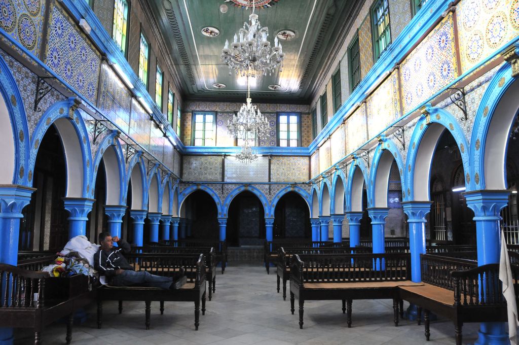 Blue columns and arches surround benches beneath a high ceiling, with patterned tiles and upper windows enclosing the hall.