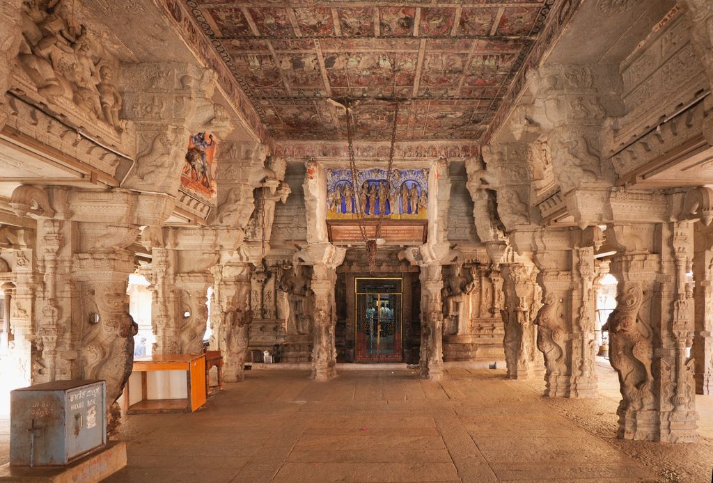 Carved stone columns frame a hall with a painted ceiling and a gated doorway at the far end; donation boxes stand to the left.