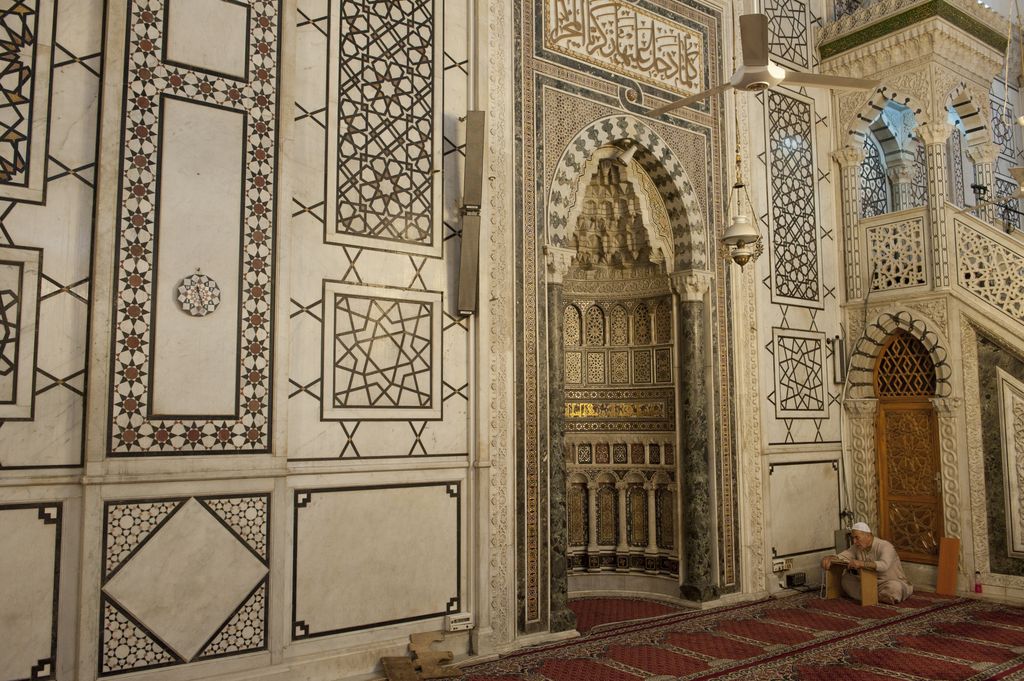 An ornamented niche is set within patterned stone wall panels beside part of a raised minbar; fans and lamps cross the foreground.