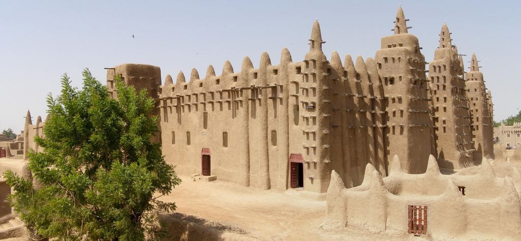 Earthen walls and three tapering minarets carry rows of projecting timber poles above a raised enclosure, partly screened by a tree.