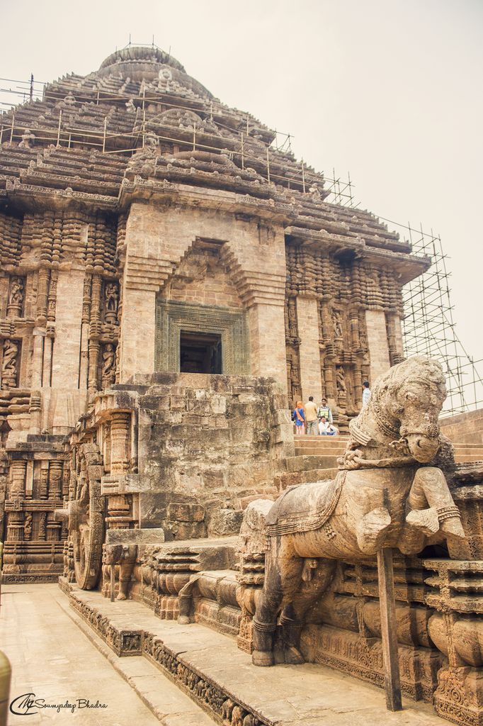 A pyramidal stone hall rises behind entrance steps, with a carved horse and wheel in the foreground and scaffolding around the upper tiers.