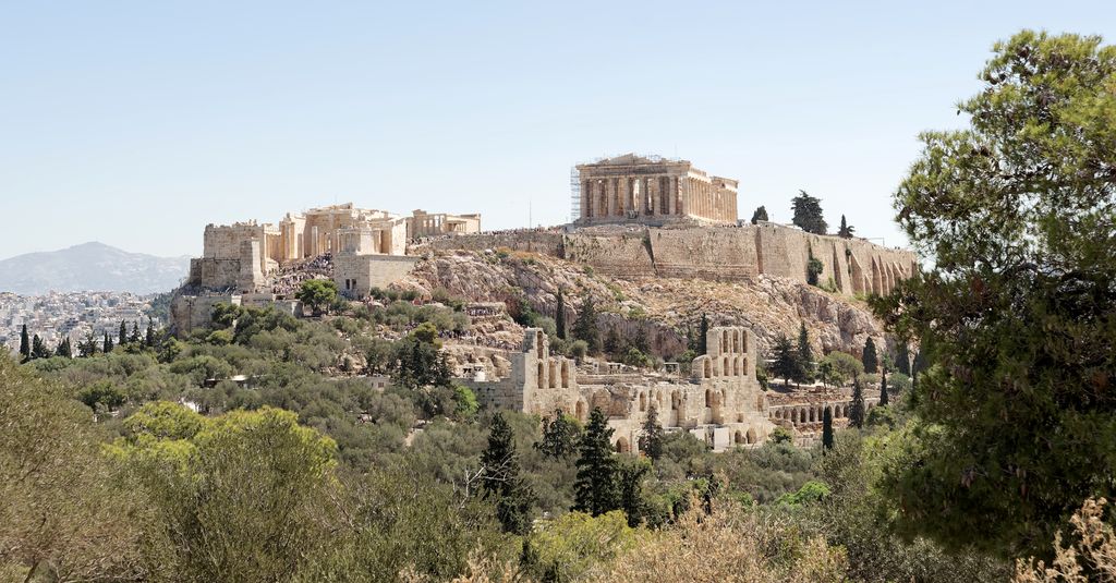 The Parthenon and western entrance of the Acropolis rise above a rocky, walled hill, with theatre ruins and trees on the lower slope.