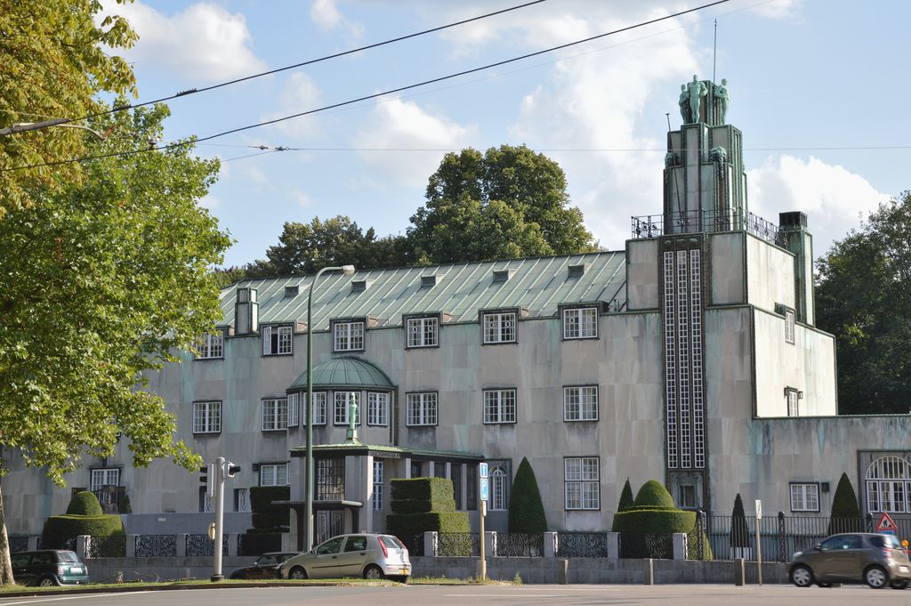 Pale planar walls, gridded windows and a tall stepped tower stand behind a street with trees, cars and overhead wires.