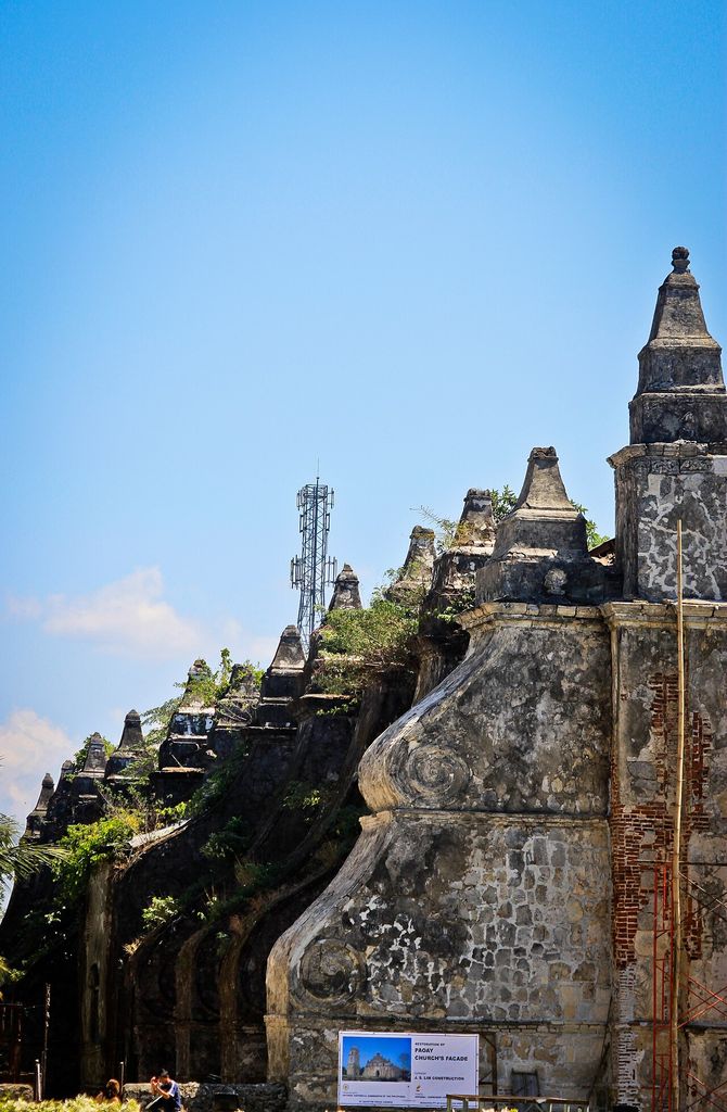 Large scroll-profile masonry buttresses project from Paoay Church, with vegetation and restoration scaffolding visible.