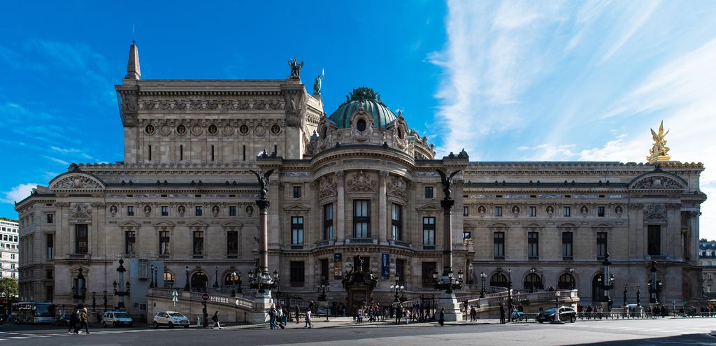 A domed pavilion projects from an ornate stone opera house with long wings, high roof masses and a curving ramped approach.