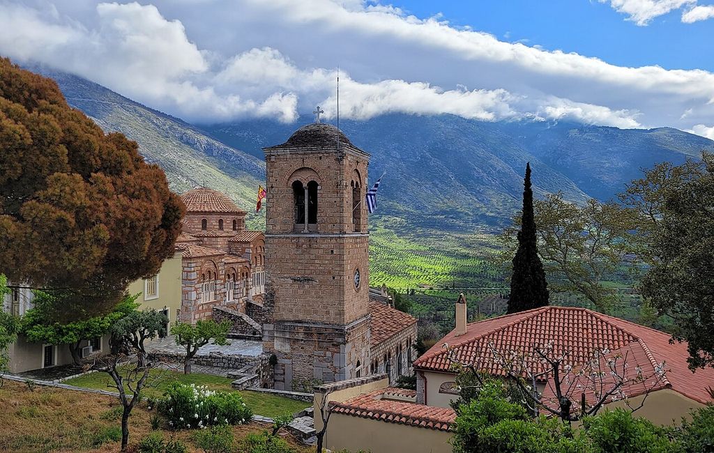 Domed church, bell tower and tiled monastery roofs beside trees, with mountains rising beyond.