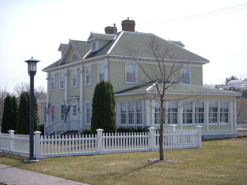 A pale yellow house repeats a pediment, tall pilasters and roof dormers, with an enclosed side wing behind a white picket fence.