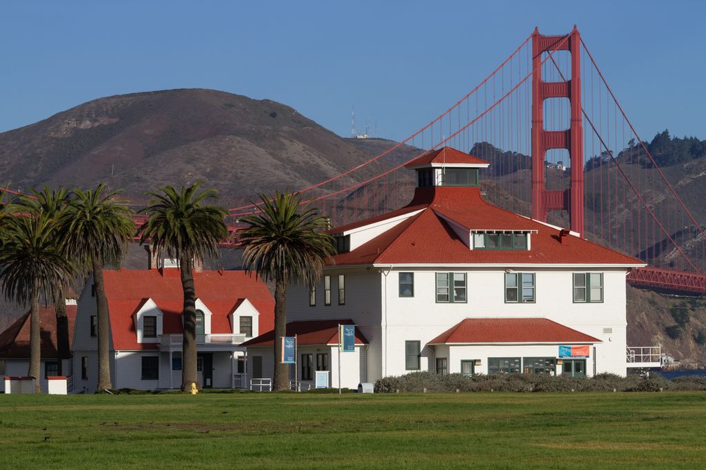 White station buildings with red roofs stand beyond a lawn and palms, with a watchtower at right and the Golden Gate Bridge behind.