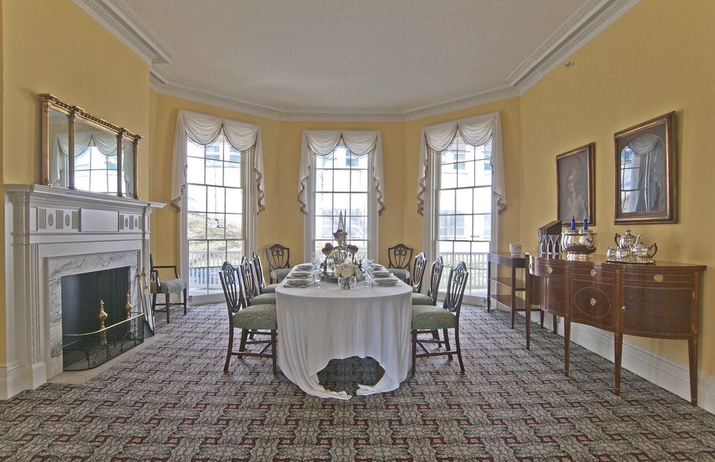 Angled wall corners and three tall windows surround a dining table beside a classical fireplace.