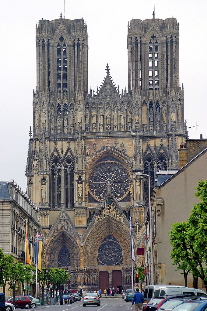 Twin towers, a large rose window and gabled entrances rise above a street lined with cars and buildings at Reims.