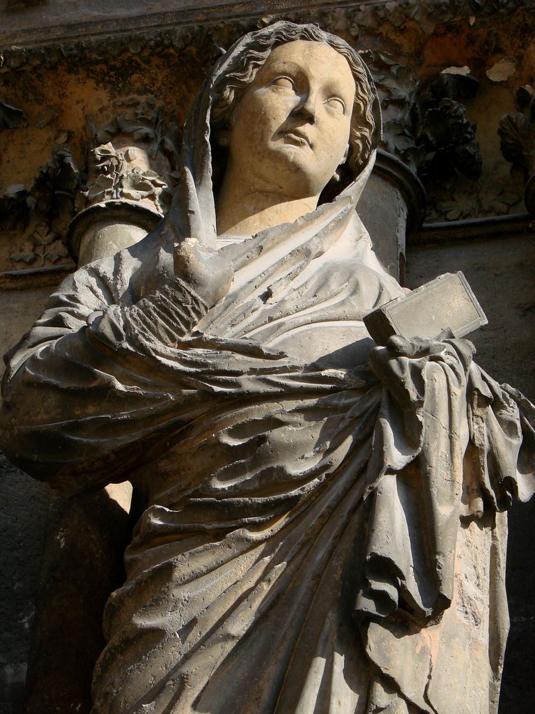 Close view of a stone Virgin holding a book, with deeply folded drapery in front of columns and foliage capitals.
