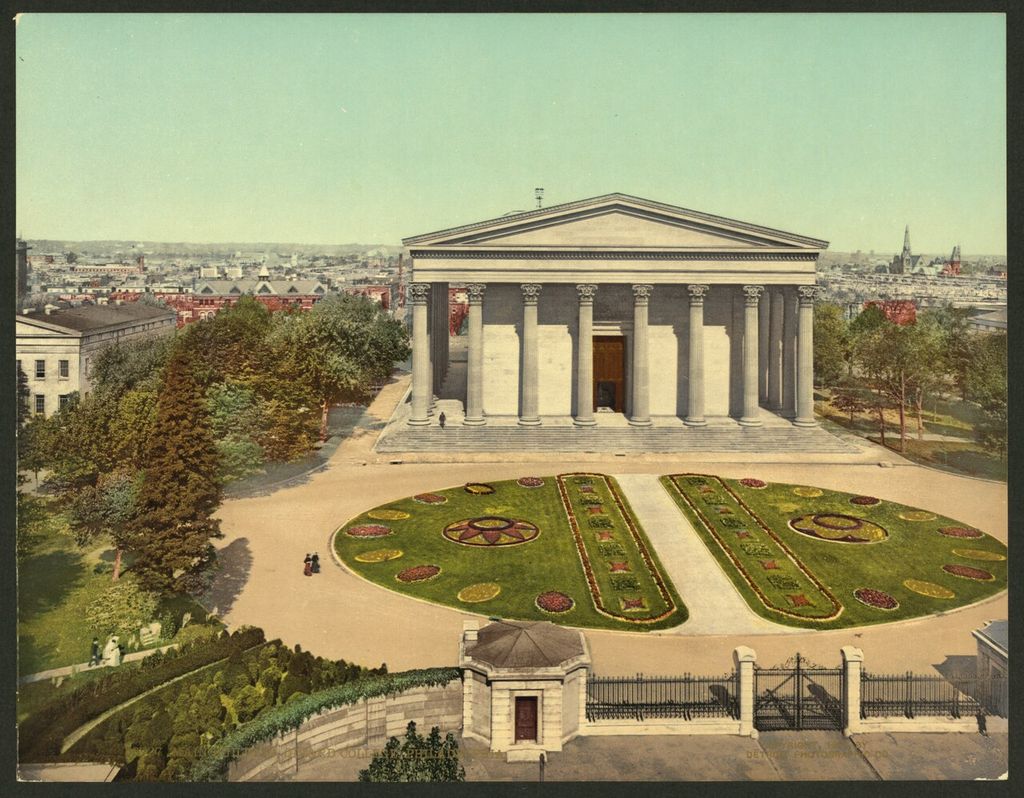 A monumental columned front rises beyond patterned garden beds and an entrance gate, with the city behind.