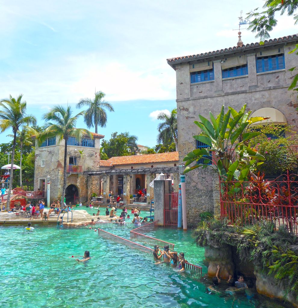 Swimmers occupy a pool beside stone-faced buildings, tiled roofs, a shaded loggia and an island linked by railings and paths.