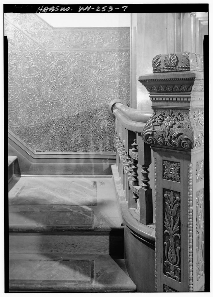 An ornate carved stair post and curved handrail stand beside an embossed scrolling wall covering in an archival black-and-white photograph.