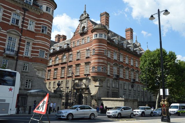A red-brick and pale-stone building with rounded corner turrets, a shaped gable and a heavy masonry base, behind road traffic.