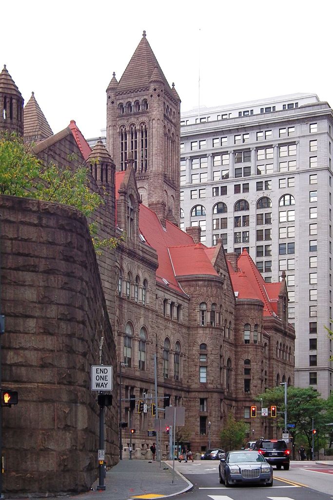 A tall masonry tower and smaller turrets rise above walls with grouped arched windows on an urban street.