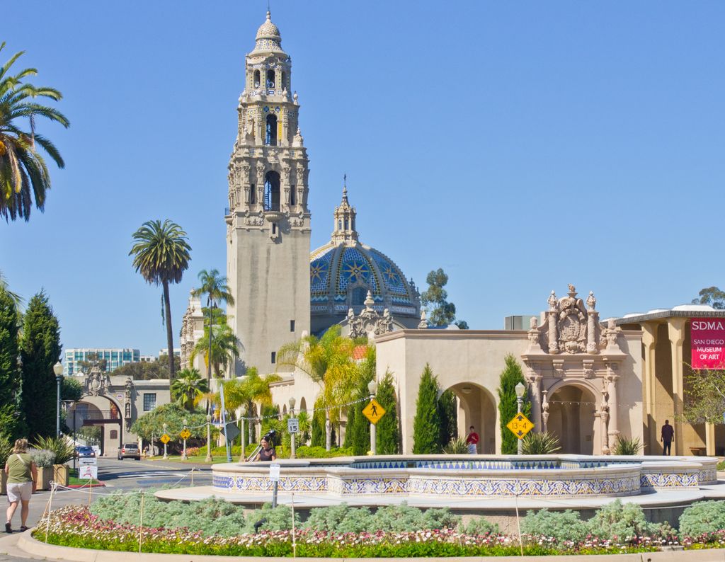 An ornate tower and blue tiled dome rise beyond palms, arcades and a tiled plaza fountain.