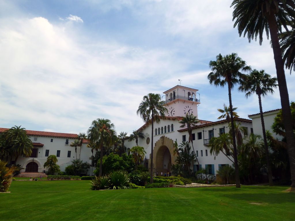 A clock tower and large arched entrance rise above white walls and red roofs around a green garden with palms.