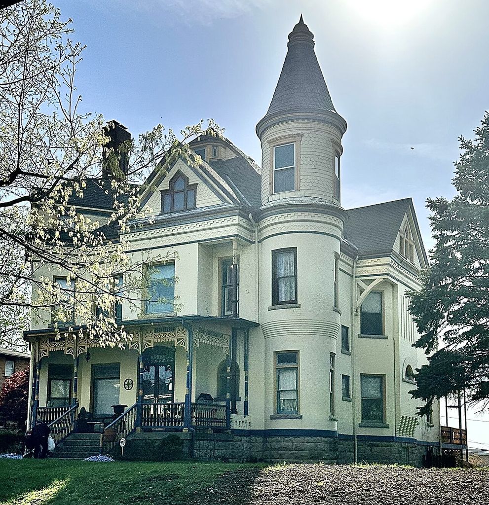 A tall shingled corner turret rises beside gabled projections and a decorated porch on an asymmetrical house.