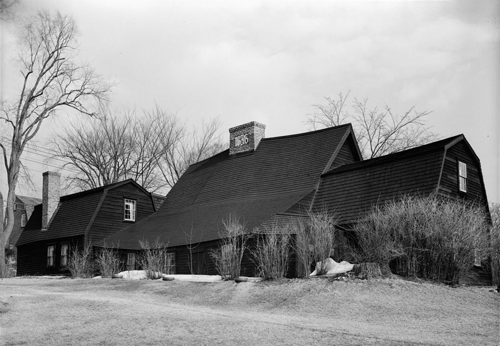 A long timber house combines a broad sloping main roof, prominent chimneys and differently roofed additions.