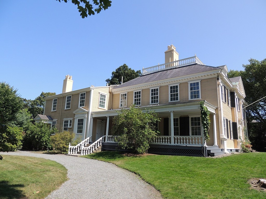A pale timber house has a hipped roof, roof balustrade, columned veranda, exterior steps and an adjoining two-storey range.