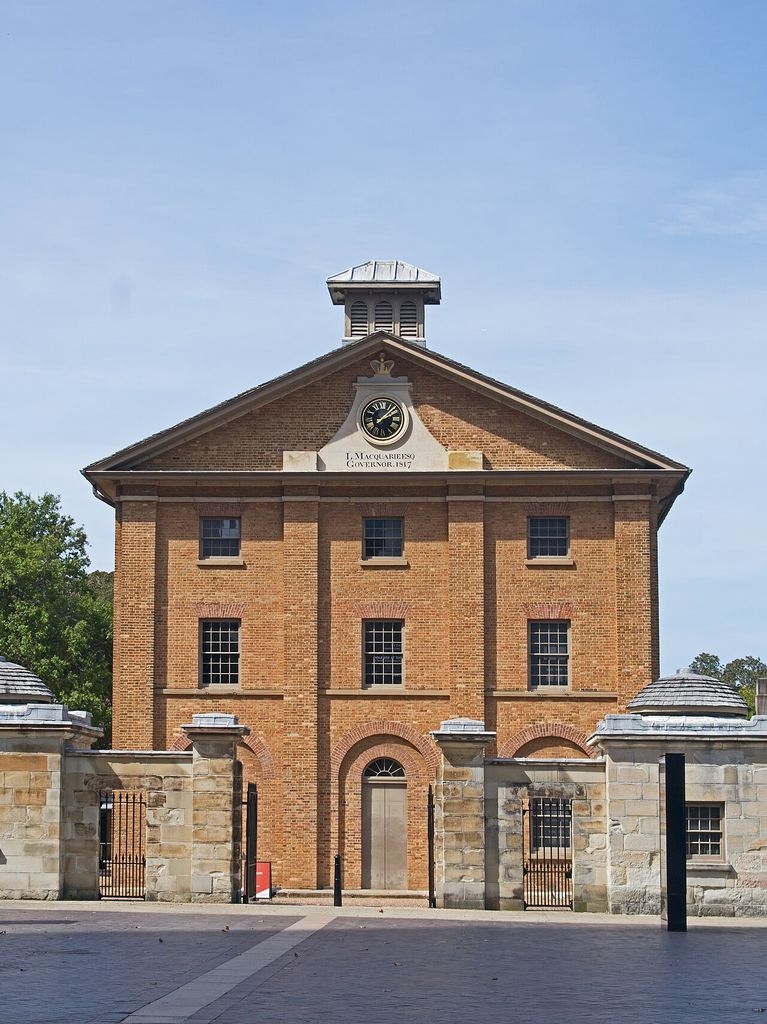 A symmetrical three-storey brick building with a clock and pediment stands behind lower stone gate structures.