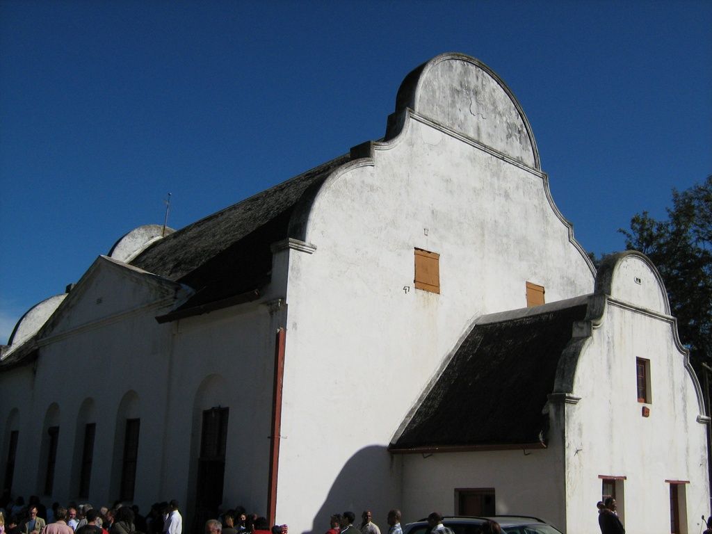 A long pale church with large curved end gables and a smaller entrance roof stands behind a gathering of people.