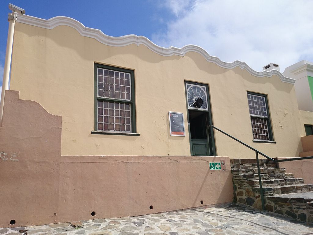 Steps rise to a central door beneath a curved parapet, with a raised stoep along the street.