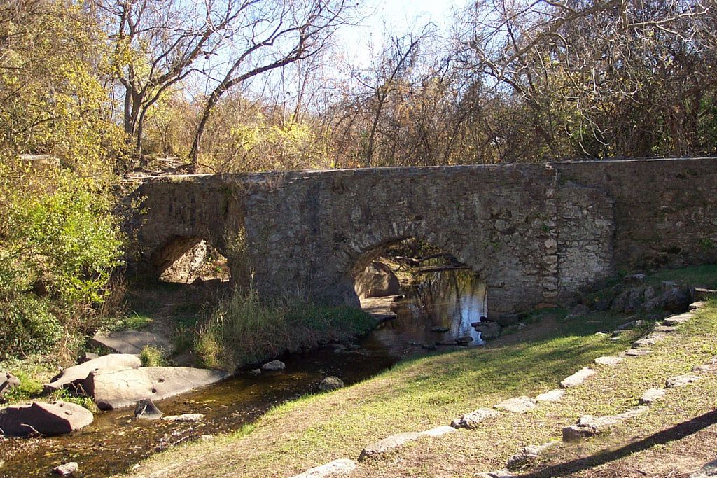 Two low stone arches support an elevated water channel above a shallow, tree-lined creek.