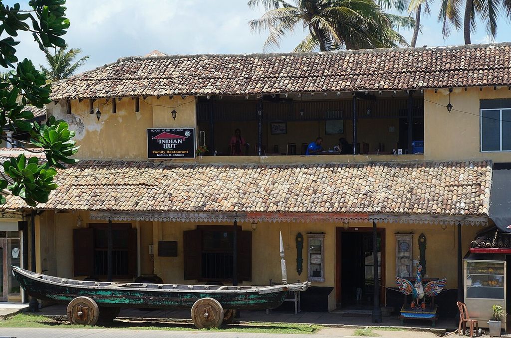 A two-storey building has a shaded upper gallery beneath broad tiled eaves, with an outdoor terrace and boat in front.