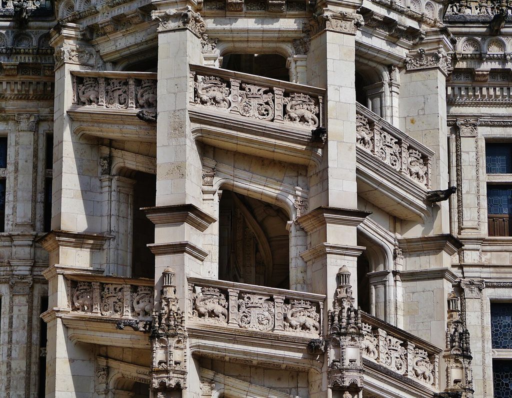 A richly carved pale stone spiral staircase projects from a courtyard wall, with open ramps, arches, thick piers and decorated balustrades.