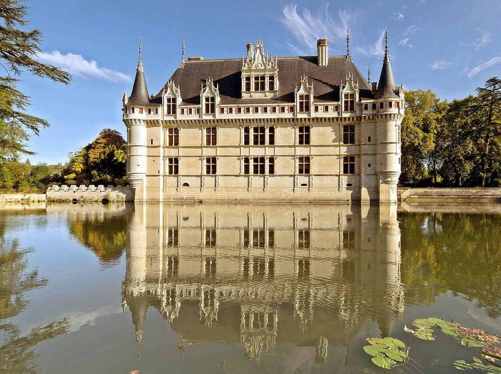 A symmetrical pale stone château with round corner turrets, steep slate roof and ornate dormers is reflected in calm water among trees.