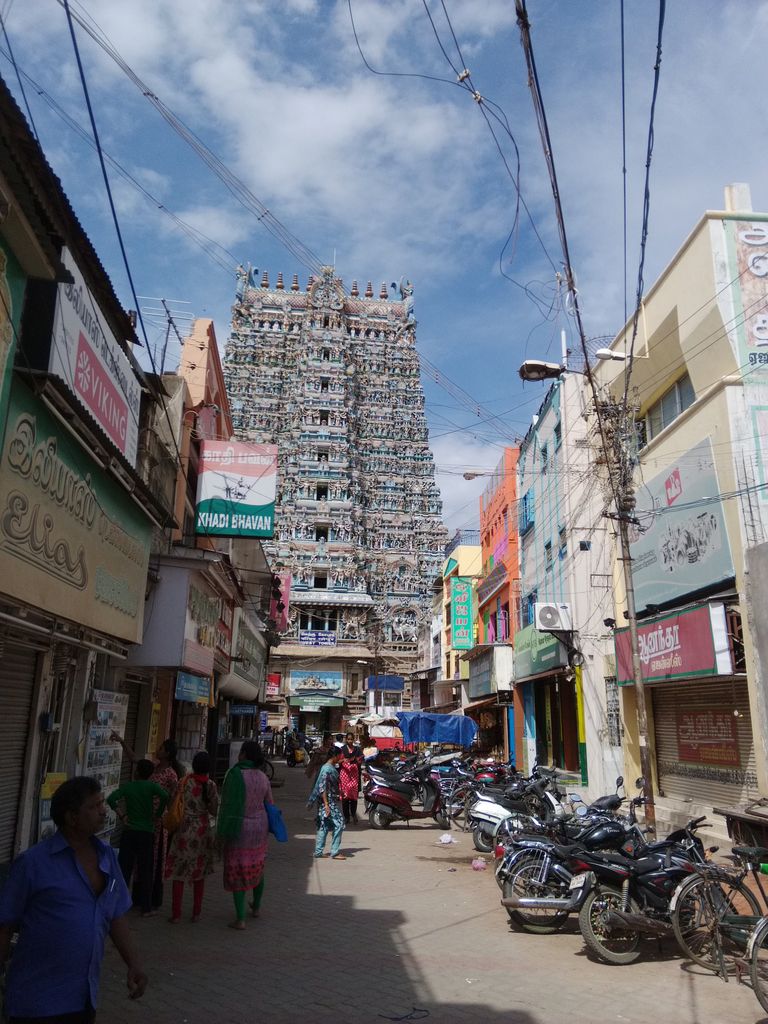 A busy shop-lined street with pedestrians, motorcycles, signs and wires leads toward a distant sculpted gateway tower.