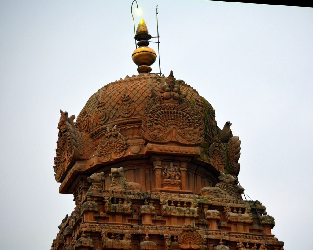 Carved reddish stone tiers rise to a rounded upper element and gold-toned finial against an overcast sky.