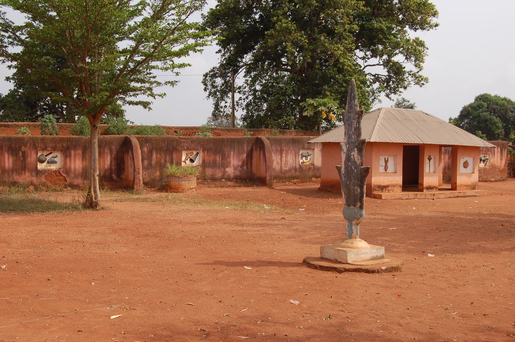 A red-earth court contains low ochre and rust-coloured walls with relief panels, a small roofed building, trees and a tall sculptural form.