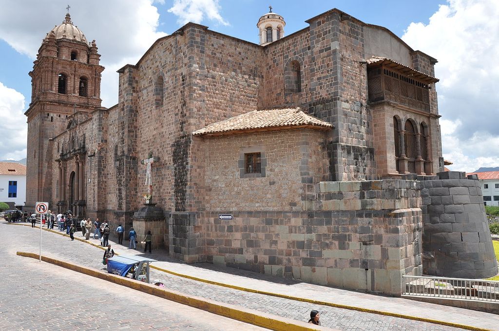 A large historic church and tower rise above coursed dark stone foundation walls along a busy paved street in Cusco.