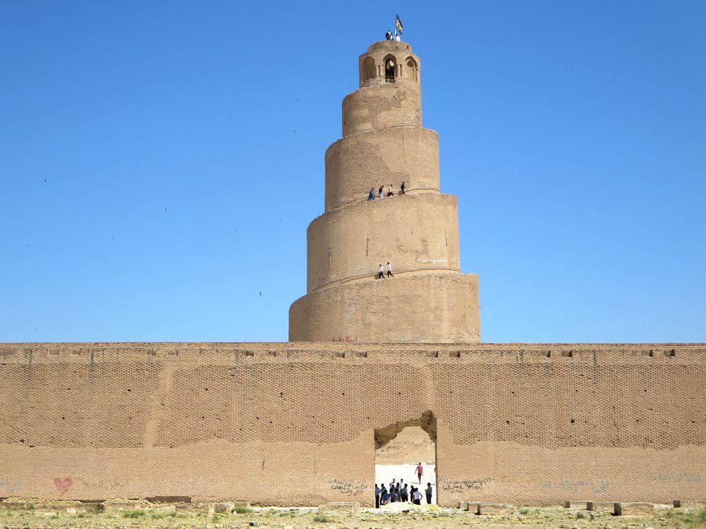 A broad freestanding spiral brick tower rises behind a low mosque wall with a central doorway and small visitors.