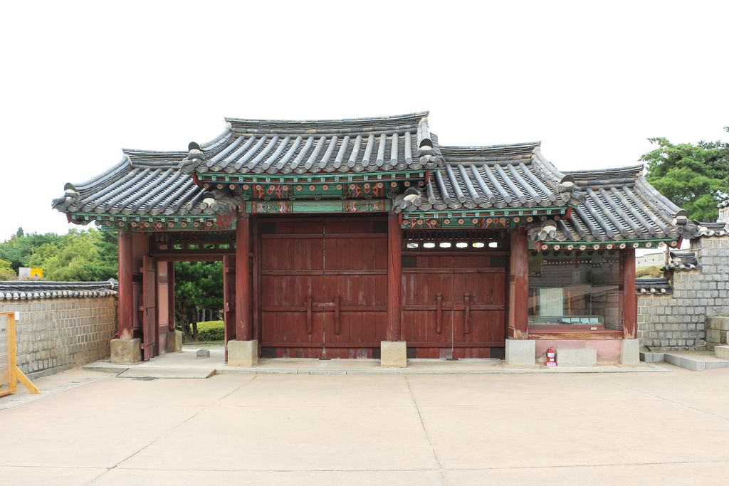 A red timber gatehouse with grey tiled roofs and painted brackets stands above pale stone bases beside a fortress wall.