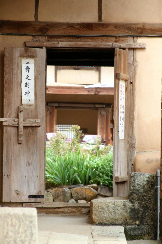 A weathered timber doorway in an earthen wall frames another gate, stone edging and green plants beyond.