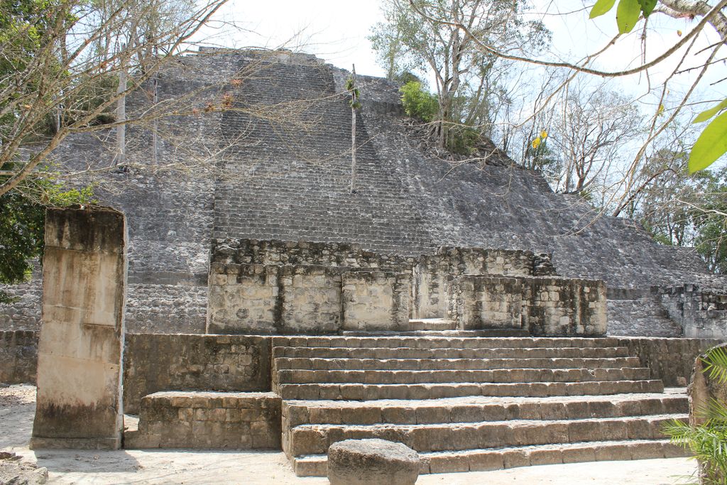 A broad weathered stone stair and stepped pyramid rise among dense trees, with a tall plain stone monument in the foreground.