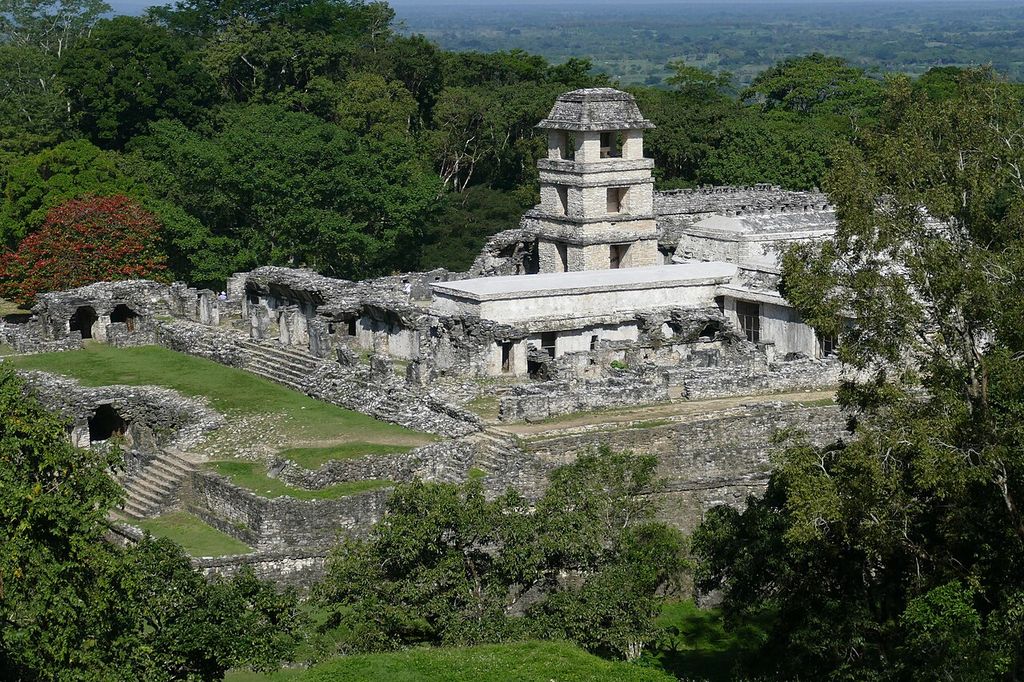 An elevated view looks across linked stone courtyards and ranges toward a square four-level tower, all surrounded by dense green forest.