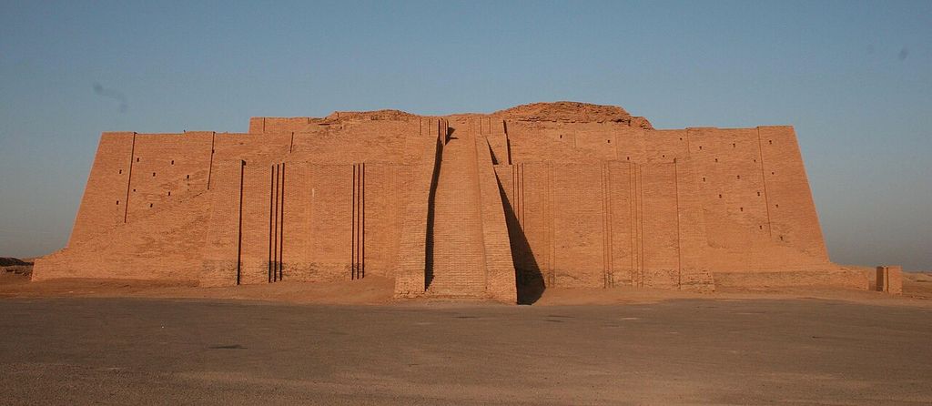 A broad reddish brick ziggurat with sloping buttressed walls and a central staircase stands against a clear sky on an open plain.