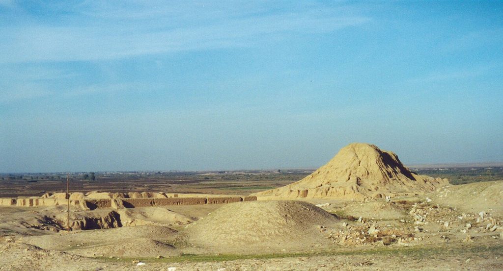 Low eroded walls and pale earth mounds spread across a dry archaeological landscape, with a larger mound and green cultivated plain beyond.
